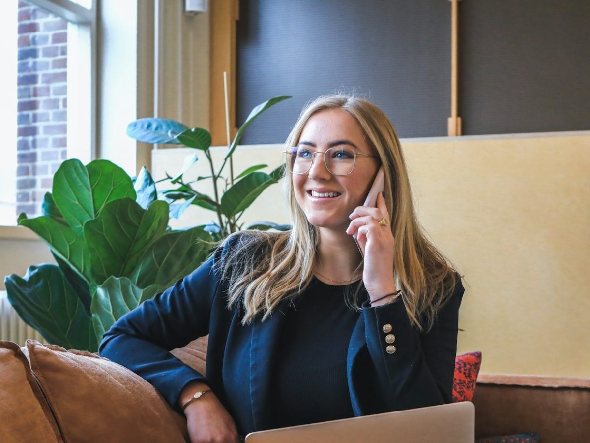 Frau telefoniert lächelnd auf dem Sofa, Laptop auf dem Schoß – Arbeiten von zu Hause